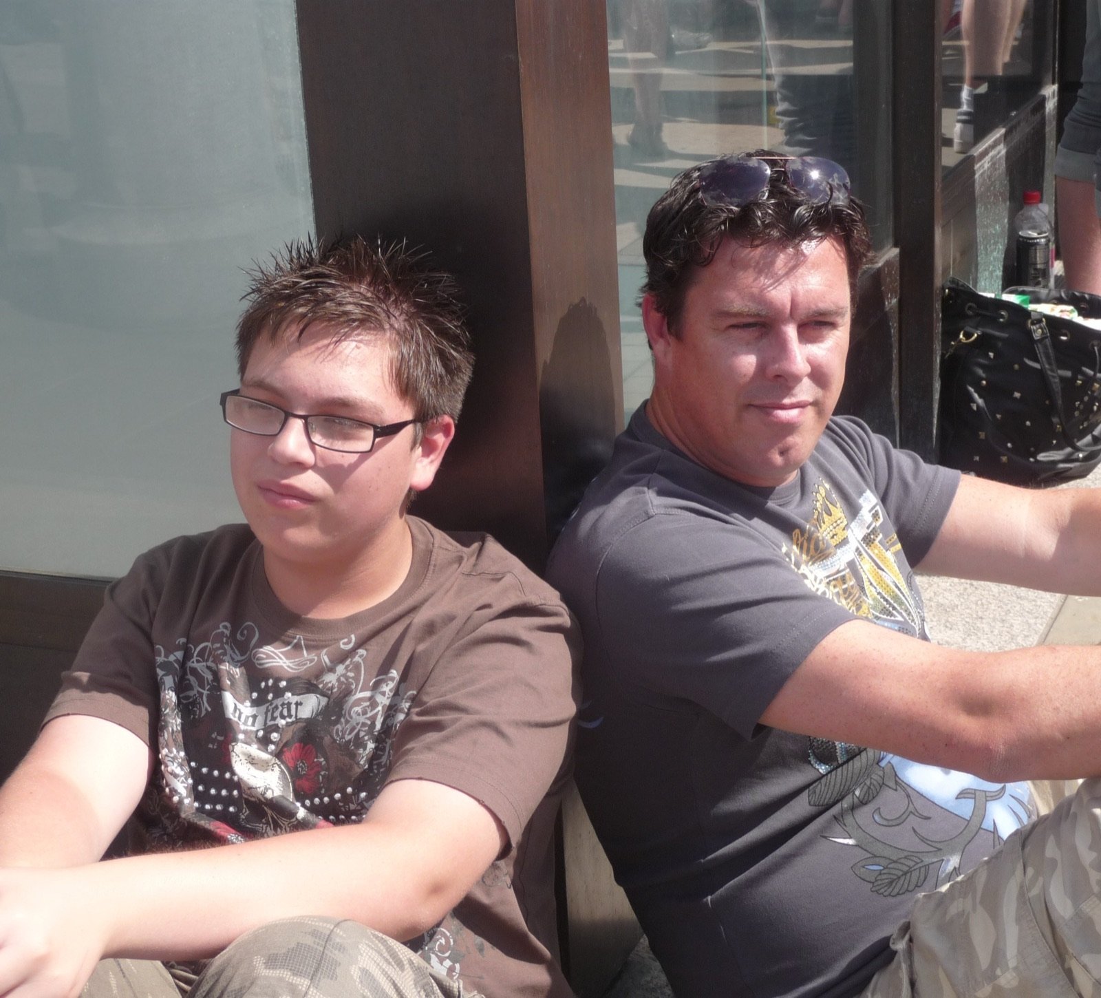 Carl and his son James sitting side by side against a shop window on a sunny day